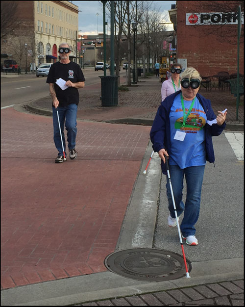 Photo shows 3 people wearing vision simulators and using a cane while crossing a street with a stop sign.