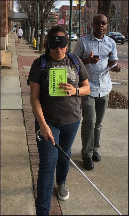 Photo shows a man and woman walking along a downtown sidewalk.  He is holding instructions for the Blindfold Walk, she is wearing a blindfold and using a cane, carrying a large card saying 'please help me to cross street, tap me if you can help.  I am deaf and blind.'