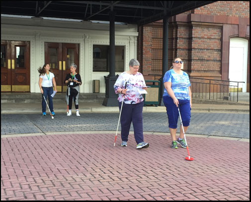 Photo shows 4 women walking from the front entrance of the hotel toward the street.  Two are walking with a blindfold and using a cane, two are carrying a cane and a paper with instructions.