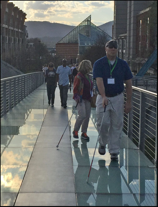 Photo shows a half-dozen people walking across a pedestrian bridge that appears to be made out of thick glass.