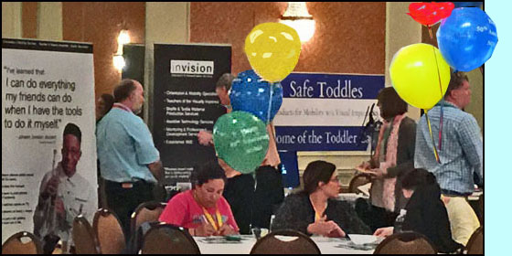 Photo shows exhibits for Invision and for Safe Toddles with people talking with the vendors.  In front of them are large round tables with balloons, and people sitting and chatting.
