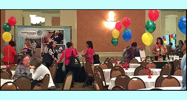7 photos all show a large room with vendors' tables along 3 of the walls, many people are at each table talking with the vendors.  In the middle of the room are large round tables with colorful balloons in the center.
