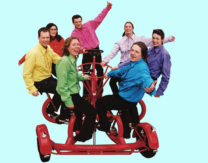 Photo shows 7 young people on the 4-wheeled red bicycle sitting in a circle and holding onto a circular bar in the middle of them, smiling at us and waving.