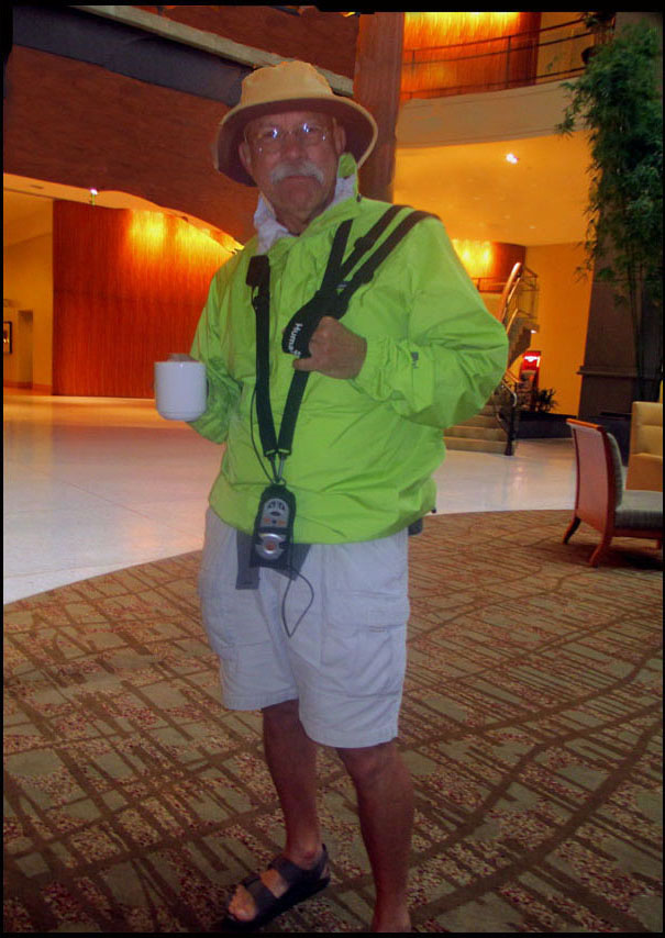 Craig is standing in the hotel lobby wearing an iridescent yellow-green windbreaker, white shorts, and a safari-type hat with a Trekker Breeze device hanging around his neck.  He is holding a coffee mug.