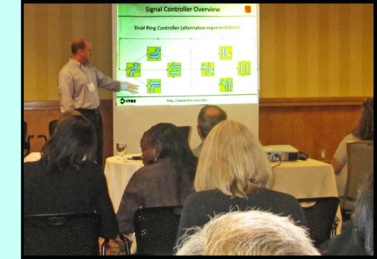 Chris Cunningham is standing on front of a room full of participants and points to diagrams on a powerpoint titled 'Signal Controler Overview.'