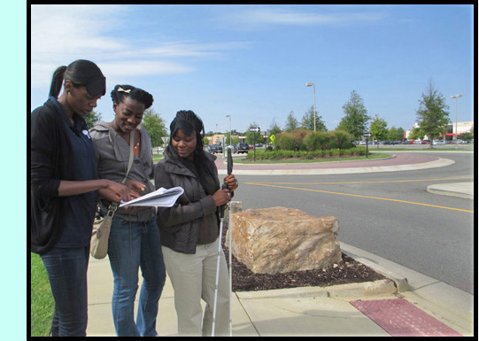 3 participants stand at a crosswalk and check their notes -- behind them we see a roundabout.